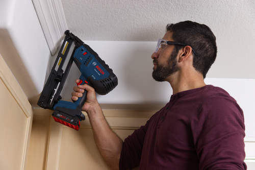 A person with a beard and safety glasses uses a Bosch GNH18V-15MA nailer to work on trim.