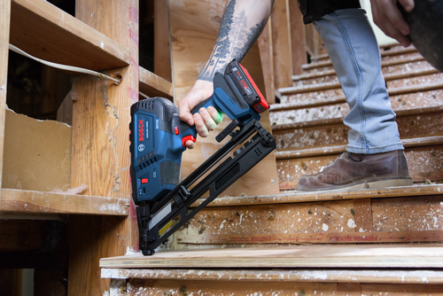Bosch GNH18V-15MA cordless nailer being held by a person with a tattoo, while standing near wooden stairs.