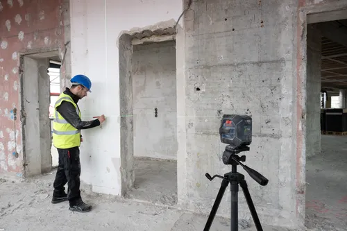 Man in a hard hat and safety vest using a Bosch laser level to measure in a concrete construction site.