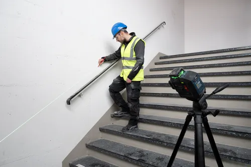 A person wearing safety gear using a laser level, with the visible laser beam aligned with the handrail on the staircase. The laser level is a Bosch.