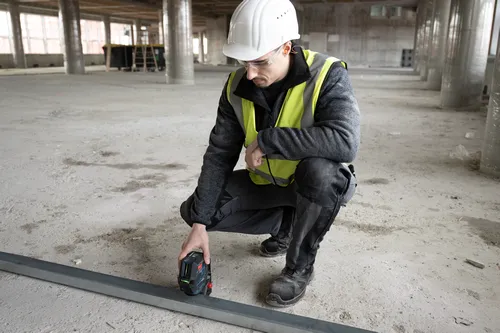 A person wearing safety glasses, a white hard hat, and a reflective vest is squatting and using a Bosch laser level on a metal bar in a construction site.