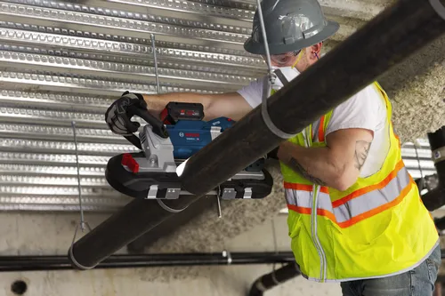 A person wearing safety glasses, ear plugs, and a hard hat uses a Bosch GCB18V-5 band saw to cut a black pipe.