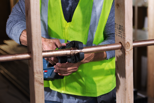 Person wearing a high visibility vest using a tool to connect copper pipes within a wooden frame.