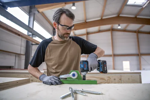 A person wearing safety glasses and earplugs uses a Bosch GDX18V-230C cordless impact driver.