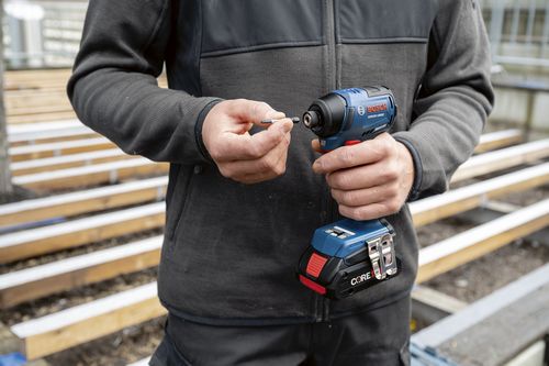 Bosch GDR18V-1950C impact driver with a bit held in the person’s hand. The driver has a battery with "CORE18V" written on it. The person wears a gray jacket.