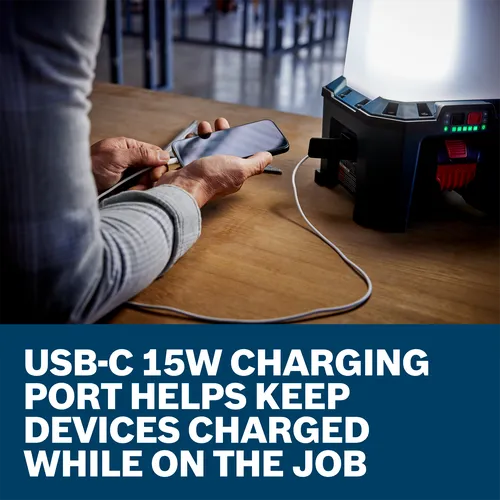 A person is holding a smartphone, connected to a USB-C 15W charging port while on the job, next to a black charging lamp.