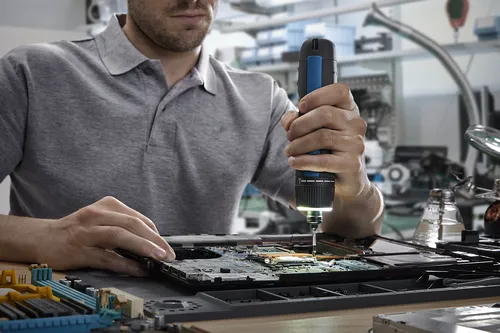 A technician using a Bosch 4V Max Screwdriver to work on a computer chip board to repair a device.