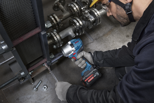 A worker using a Bosch GDS18V-780 18V Brushless Mid-Torque Impact Wrench to work on metal piping and flanges, wearing gloves and protective gear.
