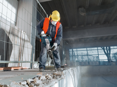 A person wearing safety gear uses a power tool to break concrete.