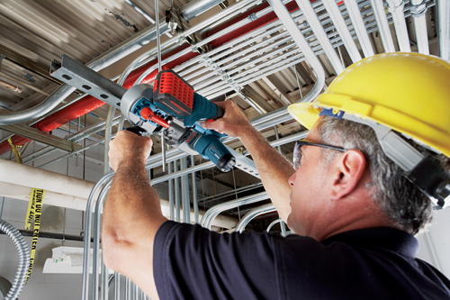 A person wearing a yellow hard hat uses a Bosch tool to cut a metal bar suspended from a ceiling with various pipes and conduits.