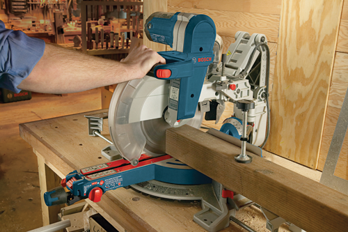 Bosch miter saw, blue and gray with visible 'BOSCH' logo, cutting a wood plank, in a woodworking workshop.