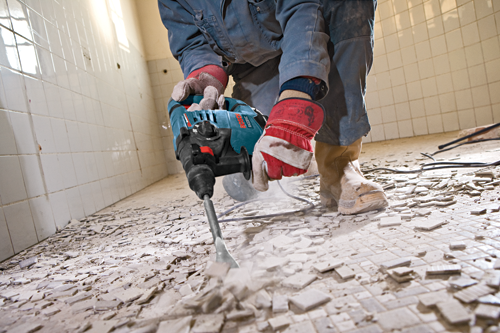 A person using a Bosch RH328VC rotary hammer to break up a tiled floor, wearing gloves, workwear, and boots.
