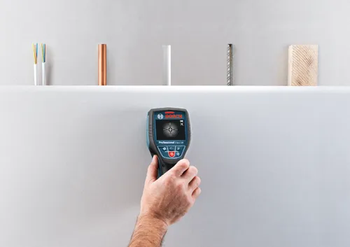 Détecteur Bosch Professional D-tect 120 tenu par une personne, détectant des objets derrière un mur, avec câblage électrique, tuyau, tige métallique et échantillon de bois en arrière-plan.