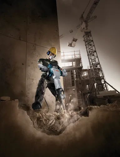 Voici la traduction :

Personne portant un équipement de sécurité utilisant un marteau perforateur Bosch pour casser du béton, avec un chantier en arrière-plan.