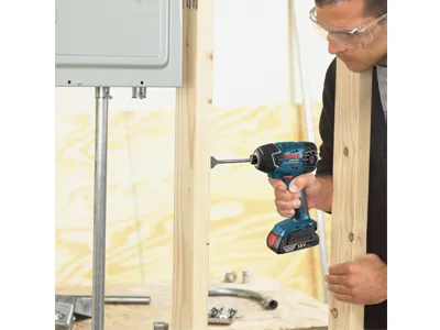 Une personne portant des lunettes de sécurité utilise une visseuse à chocs Bosch 18V pour visser une vis dans une ossature en bois.