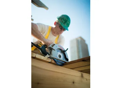 Une personne portant un casque de sécurité vert et des lunettes de soleil utilise une scie circulaire Bosch pour couper une planche de bois.