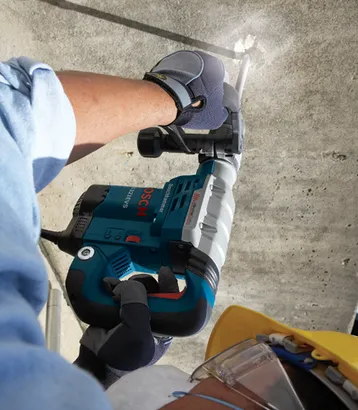 Bosch 11321EVS Boschhammer, tenu par une personne portant des gants de travail, perçant du béton. La personne porte une chemise bleue et des lunettes de sécurité. Un casque de chantier jaune est partiellement visible.