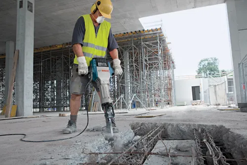 Voici la traduction :

Personne portant un casque de sécurité, un gilet de sécurité et des gants utilisant un Bosch BRUTE TURBO. L'outil électrique casse du béton sur un chantier de construction.