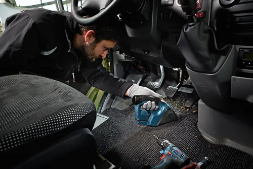Une personne portant une barbe, vêtue d'une veste et de gants noirs, nettoyant l'intérieur d'un véhicule avec un aspirateur sans fil Bosch et un tournevis sans fil Bosch.