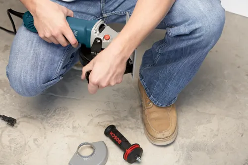 Personne portant un jean bleu tenant une meuleuse d'angle Bosch, avec une poignée noire et rouge "BOSCH" et un carter de protection gris au sol.