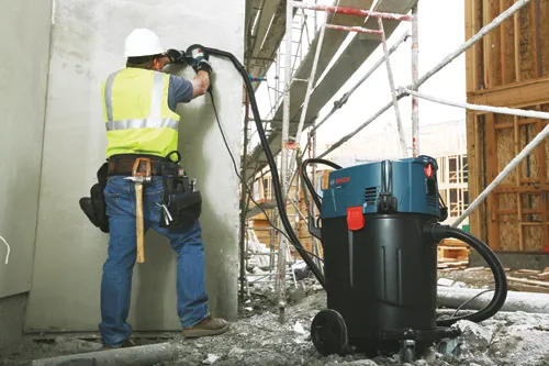 Aspirateur Bosch VAC004 avec un homme portant un gilet de sécurité et un casque utilisant un outil électrique sur un mur en béton, avec un échafaudage en arrière-plan.