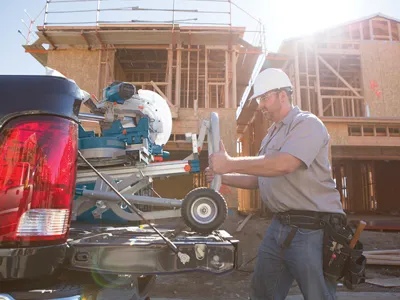 Homme portant un casque de sécurité et des lunettes de protection, debout à côté d'une benne de camion, tirant un support de scie Bosch fixé à une scie à onglet près d'un bâtiment en construction.