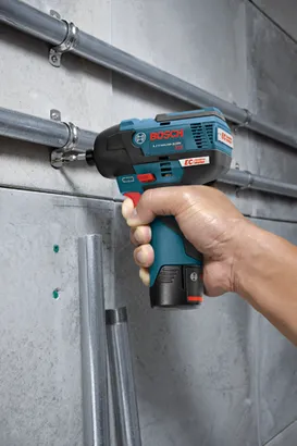 Bosch VISSEUSE À CHOC 12V EC sans balai LITHIUM-ION, tenue en main, vissant une vis dans une équerre fixée à un tuyau métallique sur un mur en béton.