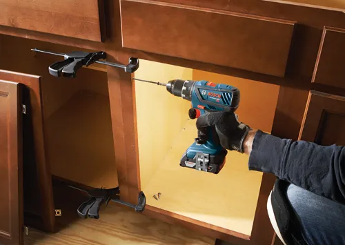 Homme utilisant une perceuse-visseuse Bosch Lithium-Ion 18V pour installer une vis dans une armoire, avec un gant noir. Deux serre-joints sont visibles.