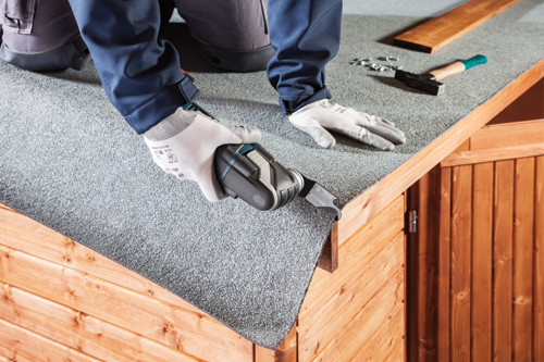 Une personne portant une tenue de travail bleue et des gants blancs coupe un matériau de toiture avec une lame Bosch ASZ 32 SC Multi Material OIS.