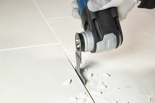 Une personne portant un gant utilise un outil multifonction pour couper le jointoiement entre des carreaux blancs.