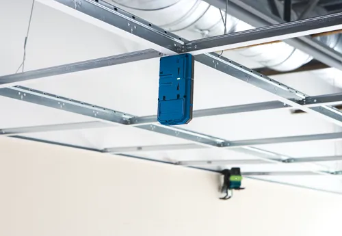 Détecteur bleu suspendu à une grille de plafond métallique, avec un voyant vert allumé.