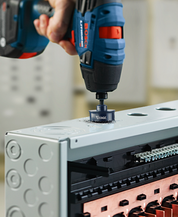 Une personne utilise une perceuse à percussion Bosch Lithium Ion pour faire un trou à l'aide d'une scie cloche Bosch sur un panneau électrique gris.