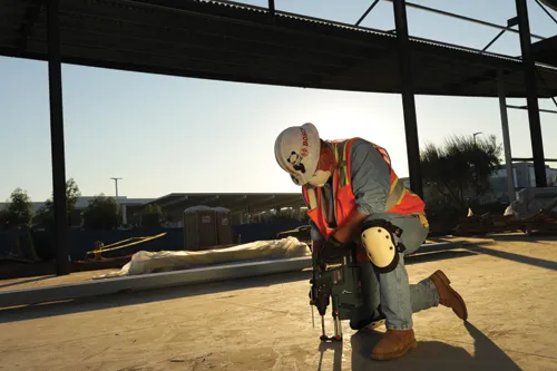Une personne, portant un casque blanc avec "BOSCH" imprimé sur le côté, un gilet de sécurité et des genouillères, perce le sol avec un marteau perforateur Bosch CORE18V sur un chantier de construction.