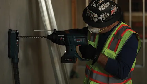 Personne utilisant un marteau perforateur rotatif Bosch GBH18V-20, portant un casque de protection, un gilet de sécurité et un masque.