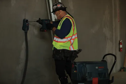 Une personne perce un mur avec une perceuse Bosch GSB18V-20, tout en étant connectée à un système d'aspiration de poussière Bosch, et portant un gilet de sécurité.