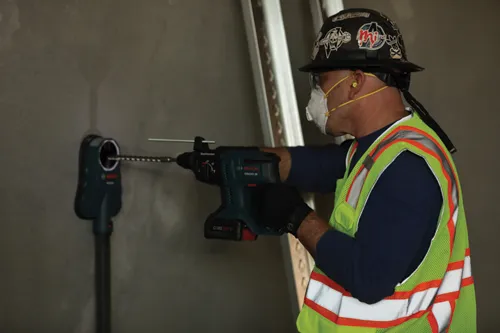 Une personne portant un casque de sécurité, des lunettes de sécurité et un gilet de sécurité utilise un perforateur burineur rotatif Bosch GBH18V-20, avec un aspirateur de poussière Bosch fixé au mur.