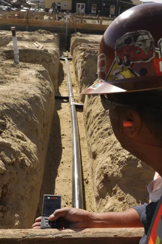 Une personne portant un casque de sécurité et des bouchons d'oreille utilise un télémètre laser Bosch sur un chantier de construction, à côté d'une conduite métallique posée dans une tranchée.