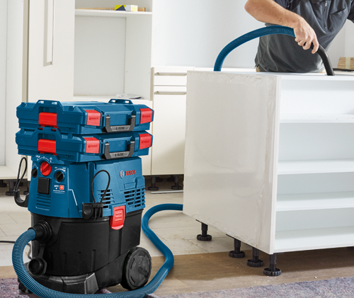 Aspirateur Bosch bleu et noir avec plusieurs coffrets de rangement rouges et bleus, raccordé à un tuyau, avec une personne tenant le tuyau, nettoyant une armoire blanche.