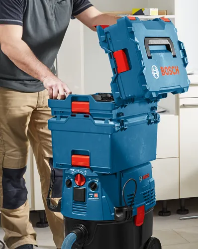 Une personne près d'un aspirateur de poussière Bosch bleu et noir avec le texte 'BOSCH' visible sur le dessus. La personne tient ouvert le dessus de l'aspirateur de poussière.