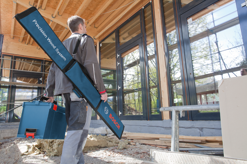 Homme portant un sac bleu Precision Wood System avec "BOSCH" visible, ainsi qu'une boîte à outils bleue, à côté d'un chantier de construction.
