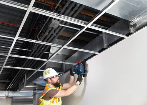 Homme portant un casque de sécurité utilisant un niveau laser Bosch avec un faisceau laser rouge sur un mur.