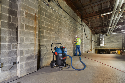 Aspirateur Bosch HEPA Ready, avec un tuyau bleu connecté à l'aspirateur, utilisé par une personne portant un casque de sécurité et un gilet de sécurité dans un bâtiment en béton, avec 2 voitures en arrière-plan.