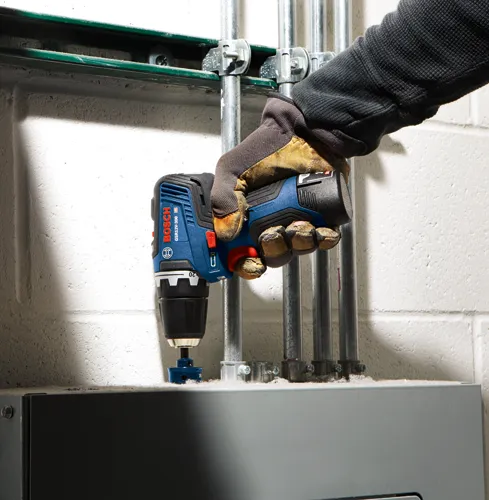 Perceuse Bosch GSR12V-300 HD tenue par une personne portant un gant et perçant une surface grise, avec des tuyaux métalliques visibles.
