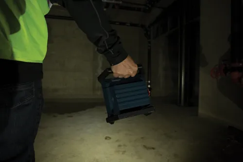 Personne portant une chemise verte et un jean, tenant une lampe portable dans une pièce en béton faiblement éclairée.