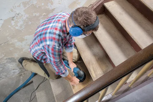 Voici la traduction :

Personne utilisant une ponceuse électrique sur des escaliers en bois, portant une protection auditive, des lunettes de sécurité et une chemise à carreaux.