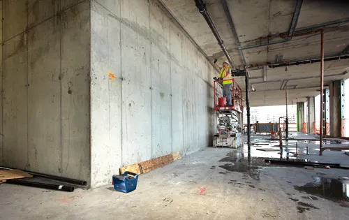 Ouvrier du bâtiment sur une nacelle travaillant sur des tuyaux ; une boîte à outils bleue est à proximité ; "134-3" et "SAFF" sont écrits sur le béton.