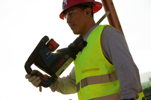 Voici la traduction :

Une personne portant des lunettes de sécurité, des bouchons d'oreille, un gilet de sécurité et un casque de protection tient une perceuse Bosch, le nom de la marque étant visible.