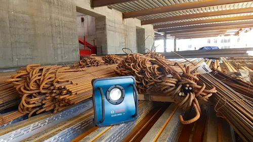 Bosch GL18V-10000C Projecteur de chantier HD sur métal ondulé, avec des tas de barres d'armature rouillées, à l'intérieur d'un bâtiment en construction.