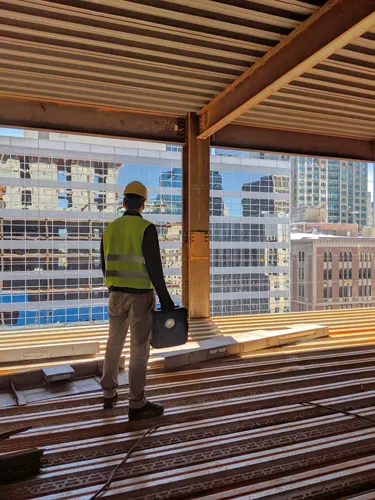 Une personne portant un casque et un gilet jaunes regarde par une fenêtre un panorama urbain, tenant un objet rectangulaire noir dans sa main gauche.