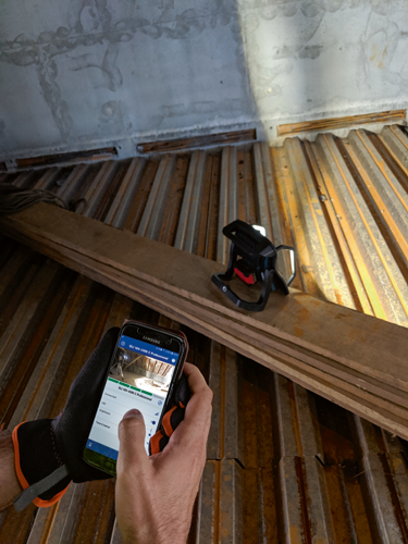 Voici la traduction du texte :

Une personne tenant un téléphone Samsung ajuste la "Luminosité" d'un projecteur professionnel GLI 18V-4000 C avec un doigt dans un environnement de travail. Une main gantée tient le téléphone, montrant le projectteur et ses commandes. Le cadre est le coin d'une pièce en construction, avec un toit métallique et des poutres en bois visibles.